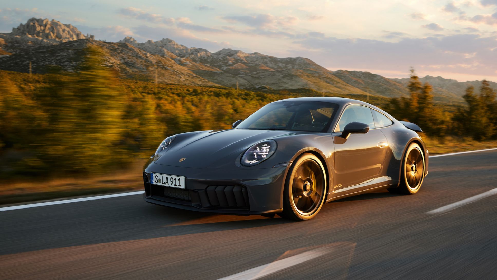 A silver 911-style Porsche with its signature sloping rear on a coastal road at sunset